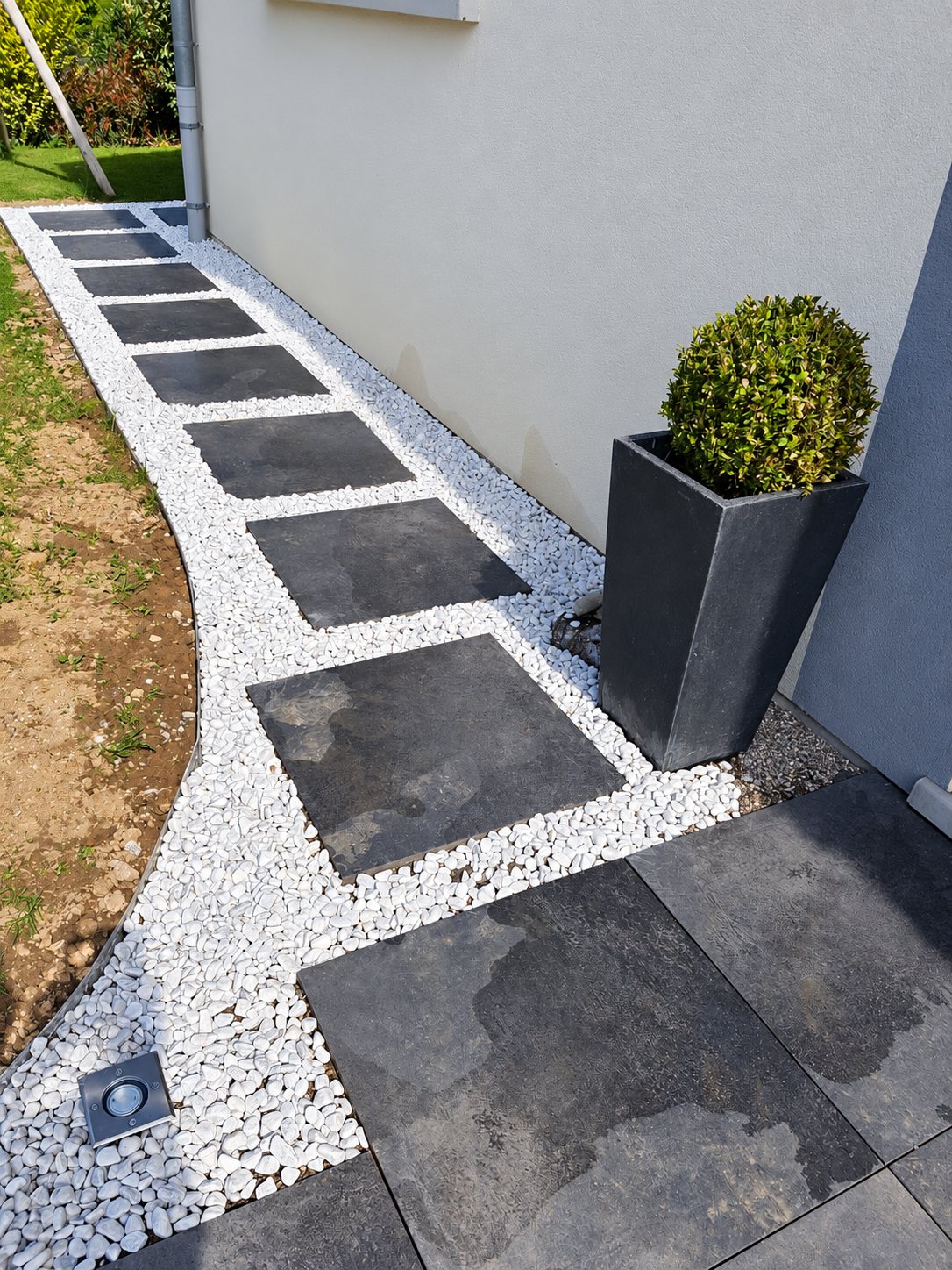 Une allée aménagée avec de grandes dalles grises foncées espacées dans un lit de galets blancs, bordée d'une bordure métallique, d'un spot encastré et d'un pot gris contenant un buis taillé en boule.
