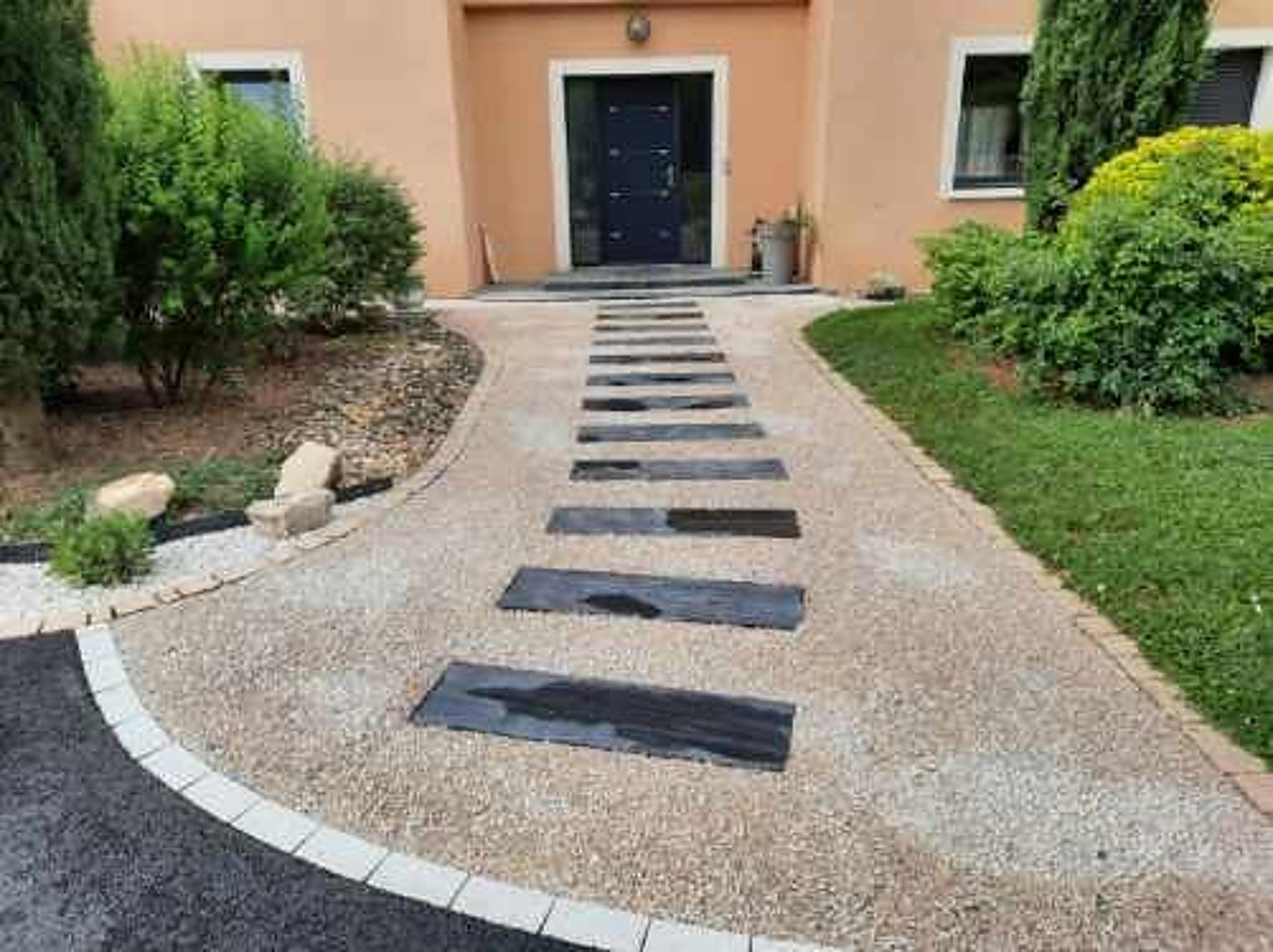 Une allée de gravier clair bordée de pavés blancs et traversée de dalles noires mène à la porte bleue d'une maison orangée entourée de verdure.