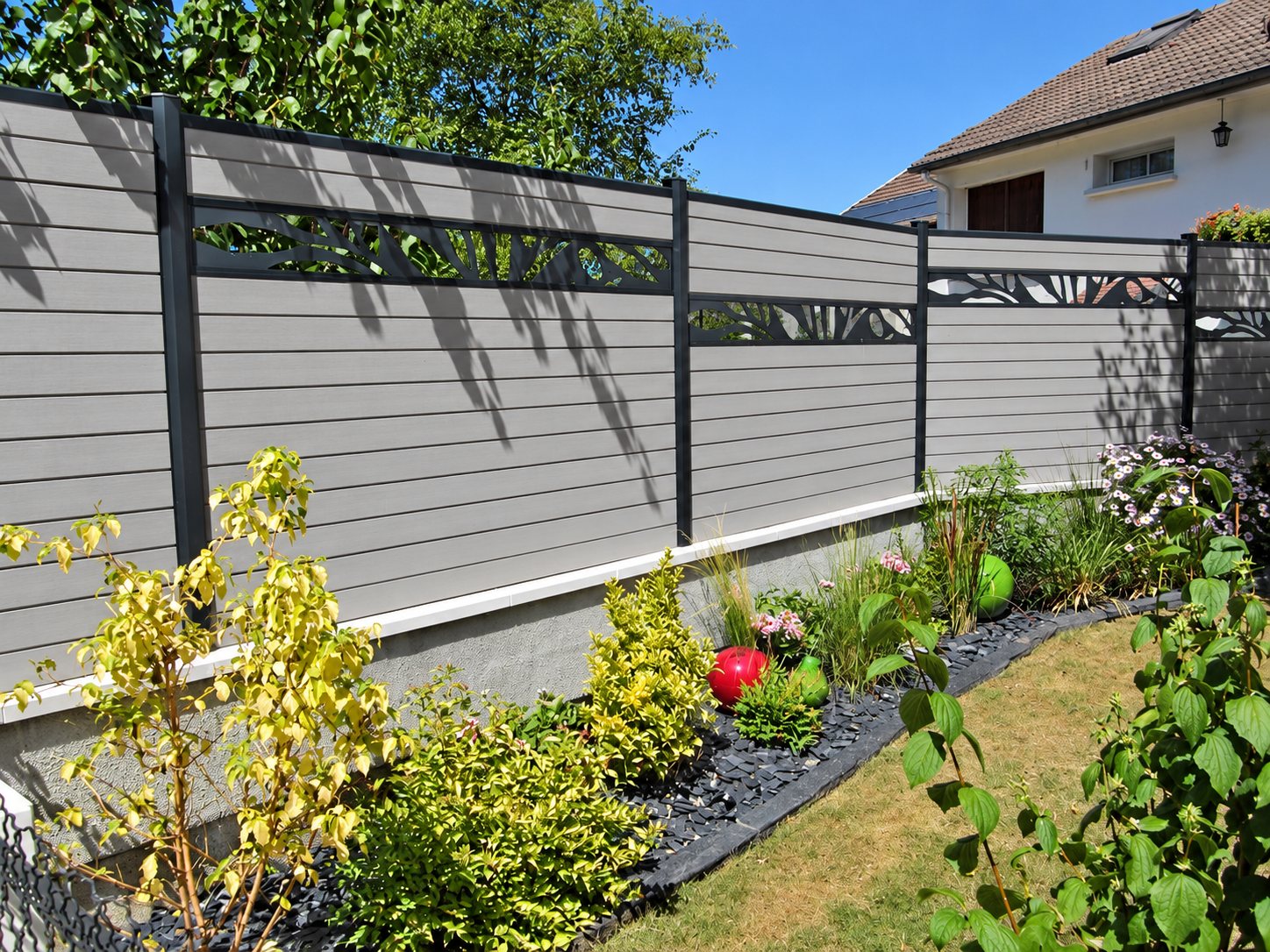 Une clôture en lames composites grises avec des panneaux ajourés décoratifs borde un jardin planté d'arbustes, de fleurs et de galets d'ardoise, devant une maison.