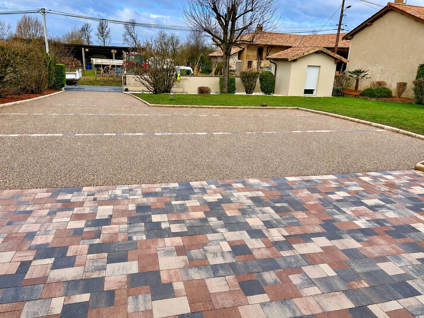 Une cour récemment aménagée avec des pavés multicolores au premier plan et un sol en gravier lavé délimité par des bordures et des marquages de places de stationnement, devant des maisons de campagne.