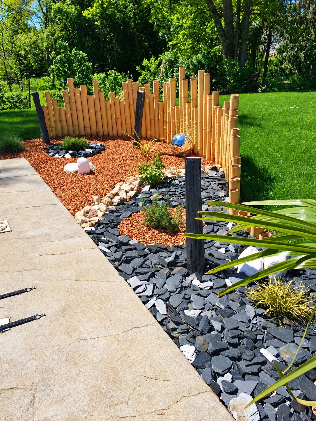 Massif de jardin paysager bordé d'une clôture en bambou, avec ardoise noire, graviers rouges, pierres blanches et une boule miroir, à côté d'une terrasse en dalle.