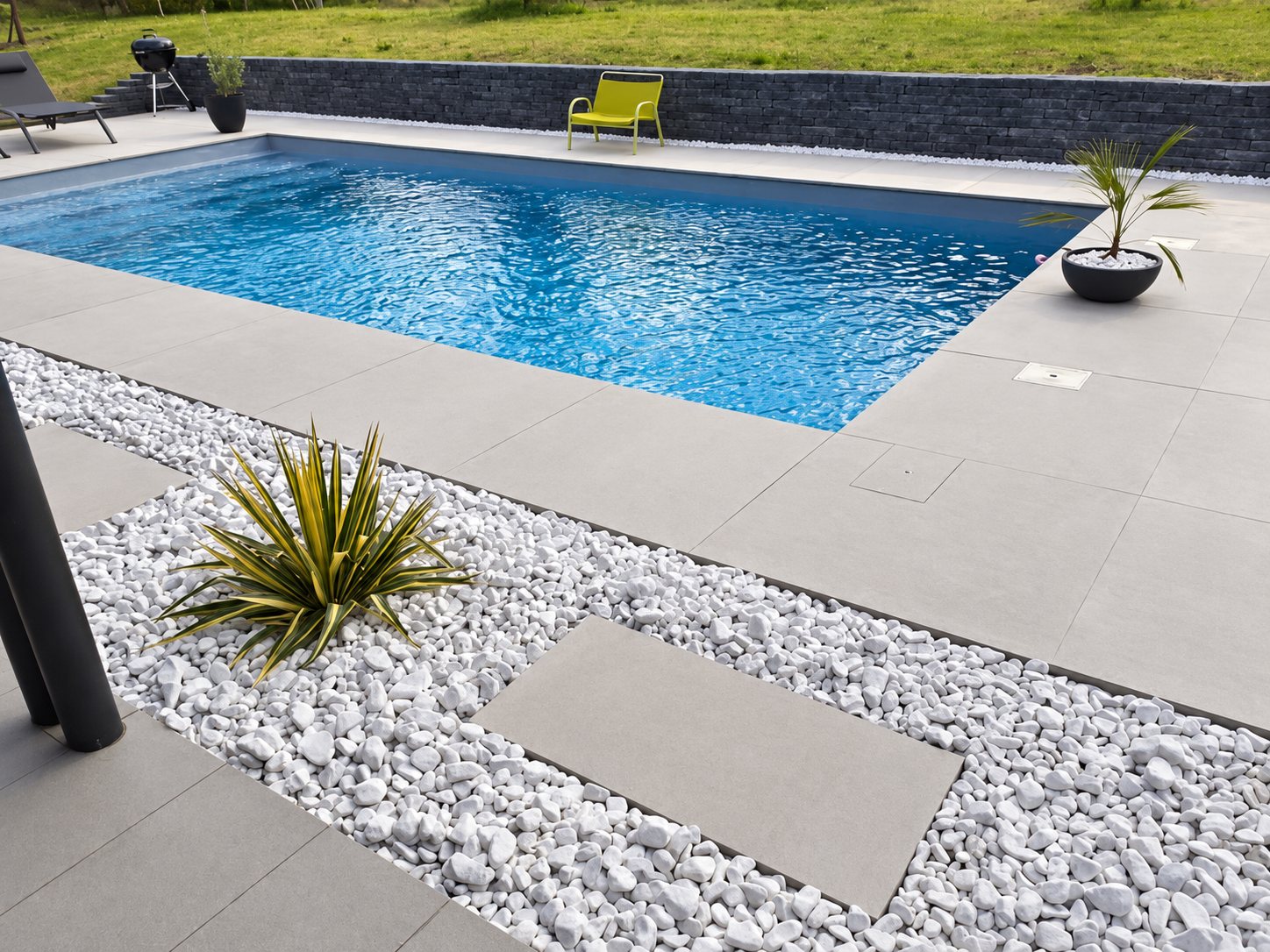 Une piscine rectangulaire à l'eau bleue est bordée de larges dalles grises et d'un lit de galets blancs avec des plantes décoratives, devant un muret en pierre sombre.